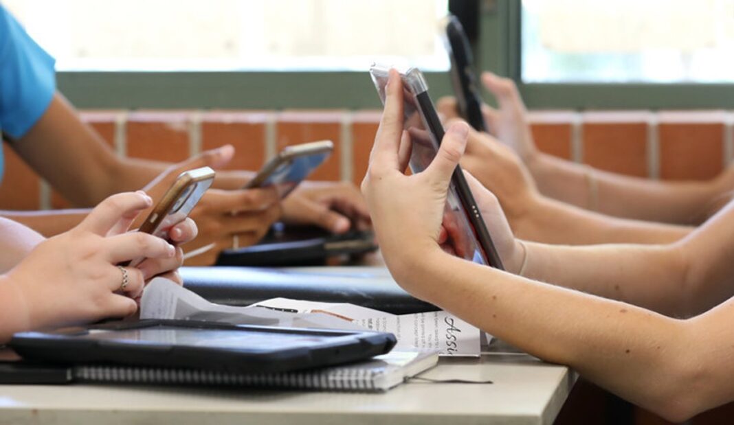 Ilustración de un celular dentro de un círculo de prohibición sobre un fondo que simula un aula escolar.